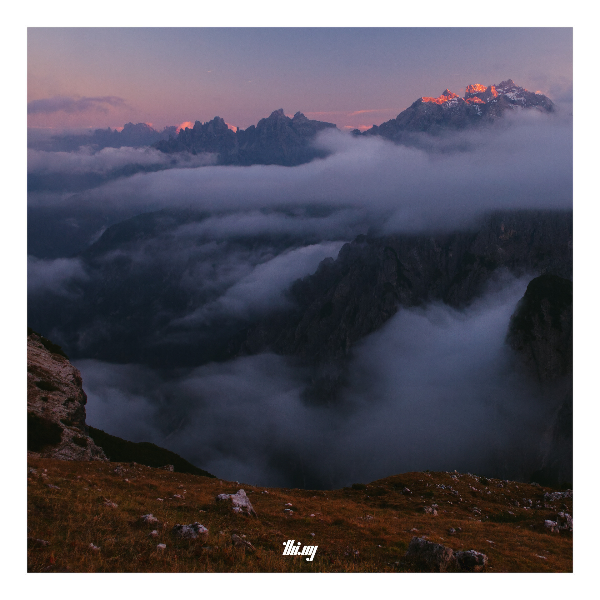 The view across and into a deep rocky valley (going down >1200m) which is quickly filling with layers of streaky clouds and fog banks drifting in, whilst the last sunbeams are illuminating the topmost parts of the jagged mountain peaks in the distance, causing intense alpenglow. The grass in the foreground, on a steep slope before the deep drop down into the valley, is tinted in rich brows/reds. The clear sky is a gradient of pinks and purples...