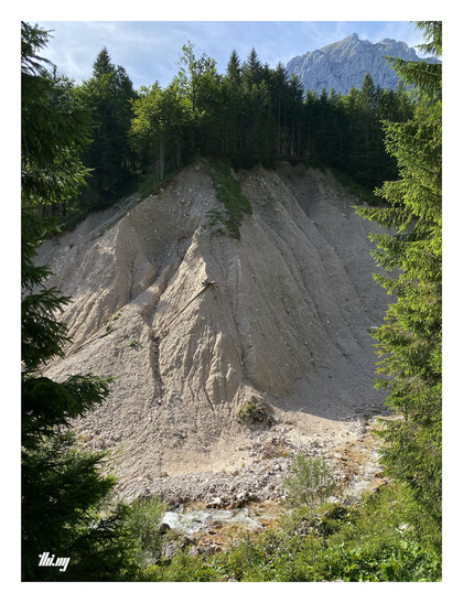 A heavily eroded steep bank of a small mountain creek flowing 20-30m below. Pale grey/beige sand with forest growing on top. Trunks of fallen trees are resting in some of the eroded creases.