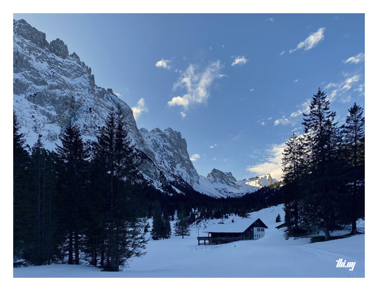 The same mountain hut in mid winter. The valley is covered in ~50cm snow. The nearby trees and forest appear almost black. The pass at the end of the valley is the only place lit by the sun, most of valley is in the shade of the surrounding mountains. Clear day with blue sky and a few isolated clouds.