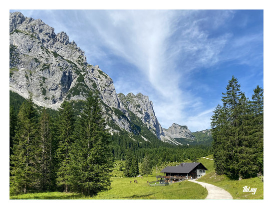 A mountain hut in a peaceful alpine valley, surrounded by meadows with grazing cows, forest and steep rock faces rising up on the LHS. The pass at the end of the valley is visible in the distance. Sunny day with blue sky and some streaky wispy clouds.