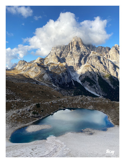 A small, shallow high alpine lake with clear royal blue water surrounded by a talus field of white sands/pebbles, located on a high plateau with autumn colored grasses in front of the large mountain massif of Crodon di San Candido. A big fluffy cloud is nestled around the mountain peaks. Otherwise beautiful blue sky.