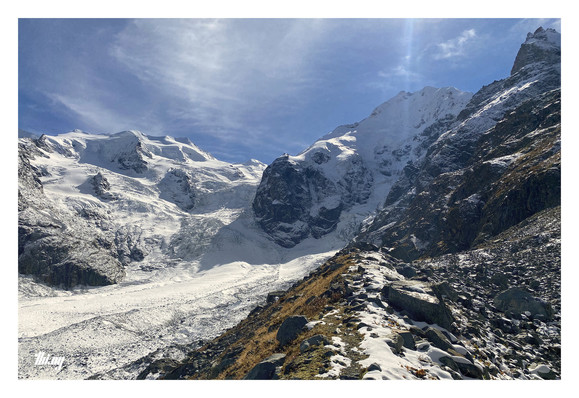 A glacier side moraine with an almost imperceptible path on its ridge is leading into a sea of ice, rocks and snow in the not so far distance. The massive glacier is just a few dozen meters below from where the picture was taken. It flows & drops down from the peaks in the back (all ~4000m and around 1300m higher up for my position), forming a maze of large seracs on the way. Sunny day with blue sky and some wispy clouds.
