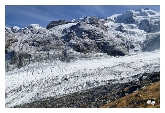 View across a large glacier (from the same moraine & position as other photo) towards the opposite side of the valley with its 4000m peaks, hidden below a thick layer of snow and ice, some side glaciers and bare major rock faces dusted in snow. Some dried brown grass in the bottom-right corner.