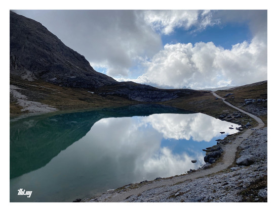 A narrow path leading along the edge of a shallow high alpine mountain lake towards the horizon. The lake has crystal clear cyan-blue water, its surface completely still and reflecting the clouds above. A steep rock face is leading up into a dark cloud on the left-hand side (only partially visible). A little blue sky in the far back.