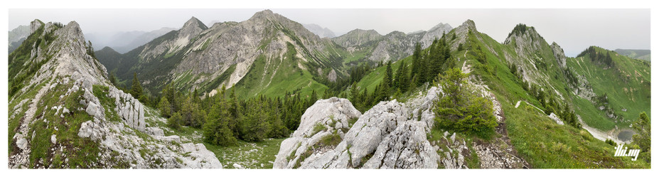 Panoramic photo of a narrow rocky ridge line with steep beautiful green grassy slopes, mountain passes and forest in nearby valleys. A small lake at the bottom of the doline (RHS in the photo). The rocks have a beautiful light gray color, almost the same as the overcast, cloudy and misty sky, heavy with humidity (80%+).