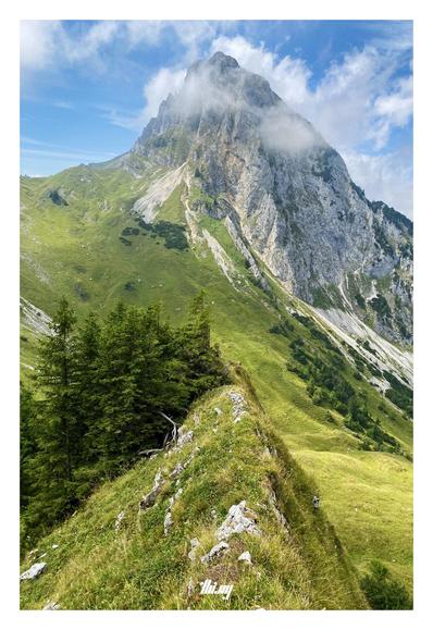 View of a mountain and nearby pass from a narrow & steep smaller ridge. The main peak is semi-shrouded in clouds and has large rugged rock faces reaching down into the valley on the right. Lush green grassy slopes are leading down from the pass. Small isolated patches of trees .