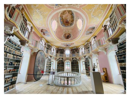 Wide-angle view of the baroque library at St. Mang monastery in Füssen: An oval room with rose/pink walls, white bay windows, floor to ceiling bookshelves filled with books from 1780s onward. A narrow gangway along the walls with carved wooden railings and angels is forming a second level. Yellow ceiling decorated with beautiful frescoes and a false cupola with additional windows to let in more light. Below it there's an small oval cut-out in the floor to view the concert room below... 