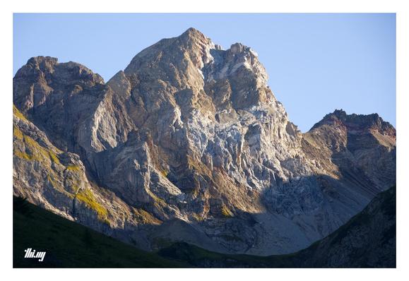 Photo of a rugged mountain peak with its main face showing layers of different sedimentary rocks, shades of white/light gray, beige, brows, deep reds. A few slopes on the side covered in grass, illuminated by the evening sun. The pass and valley below are already in the shade of the slowly setting sun. Clear, pale blue sky.
