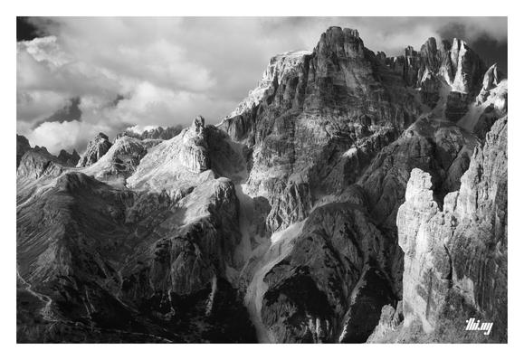 B&W photo of a majestic and rugged mountain massif on the other side of a very steep valley. The view is from the ridge/pass. The vertical rock faces/needles in the foreground and some parts further back are illuminated by the autumn afternoon sun. The rest is in semi-shade cast by a layer of clouds moving in. Some parts of the massif are showing distinct traces of former glacial activity. A grandiose view!