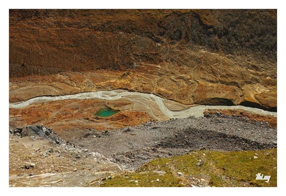 Top down view of the Langtauferer outflow section of the valley, showing a patch work of different textures/rocks and colors from grassy slopes, talus fields in shades of gray, orange, beige, rust. The milky grey glacier creek meandering through it all from left to right. A small pool of crystal clear intensely green-blue water nearby. The entire scene feels like an abstract painting