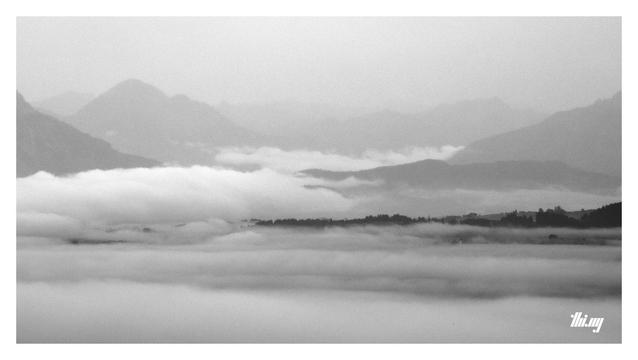 Black and white photo looking into the Lech valley near Reutte (in the distance), from above layers of fog and low hanging clouds. Silhouettes of forest and mountains fading into the gray background.