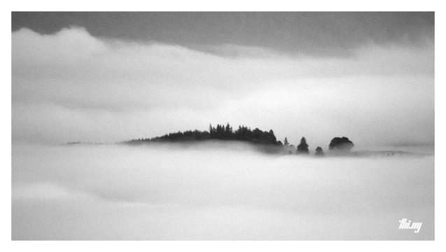 Black and white photo of the silhouette of a  hill top surrounded by dense layers of fog.