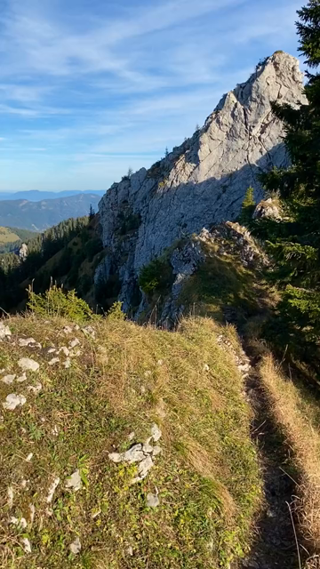 First person video, walking on a narrow path along a ridge, looking down very steep slopes on either side, covered in dried grass and forests, rocky cliffs. Views as far as the eye can see. Late afternoon sun with beautiful warm light. Serene clear sky.