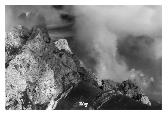 B&W photo of a craggy rocky mountain peak with several steep cliffs and a smooth grassy mountain pass curving further down on the right. Dramatic sky with clouds moving through the peaks and larger cumulus building up in the background.