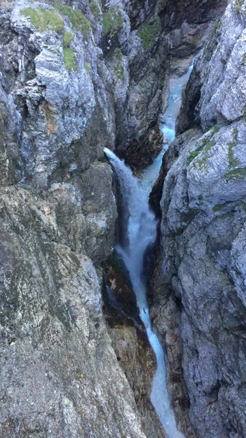 A karst spring cum waterfall gushing from the wall inside a steep and very narrow canyon, viewed from a small bridge ~60m above. The camera then slowly pans upwards, revealing the full vertical depth of the canyon and surrounding valley...