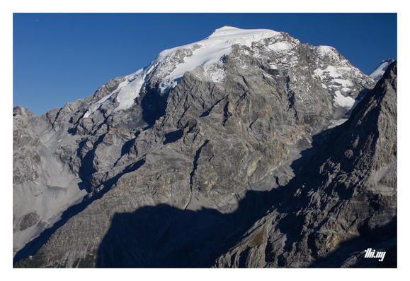 View of the Ortler mountain with its glaciated peak and prominent nortwest ridge leading down deep into the valley. A tiny speck — actually the 2-storey mountain hut (Berglhütte) — is near the (visible) bottom of that ridge, just above the tree line. Another glacier (Niederer Ortlerferner) was coming down in the valley in front of the ridge, but by now stops much further up, its tongue barely visible in the shade... Deep blue, clear sky.