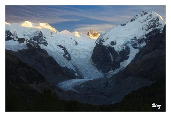 View of the grand Morteratsch glacier, its valley and surrounding peaks of the Bernina range (incl. its namesake Piz Bernina, 4050m, on the right). Most of the valley is already in deep shade, only the highest peaks are illuminated in golden sunset light. On the right side of the valley, the line between vegetation and gray rock in the parts where the glacier has already retracted, marks the previous extent of the glacier. Large pieces of exposed bedrock. Blue sky with streaky clouds. 