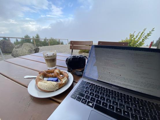 Photo of a garden table with laptop, camera, brezel and coffee (at a summit restaurant). Some tree tops behind the falloff and clouds behind that. Intermittently a few gaps of blue sky.