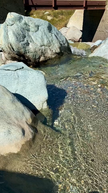Short video of a crystal clear mountain creek with a small rusty footbridge, the camera panning around showing a high alpine landscape with glaciers, rock faces and the higher up parts of the creek, filled with boulders and a small waterfall
