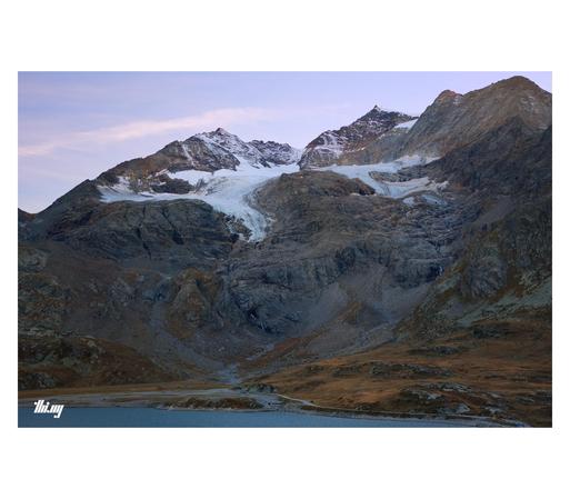 Photo of a glaciated mountain massif (Piz Cambrena) in a vast high alpine landscape. The two main branches of its glacier have largely retracted and only the higher up parts are now just feeding several waterfalls flowing through the exposed bedrock further below. Steep bare rock slopes, giving way to rolling meadows of brown dried grass near the pass. A sliver of the large mountain lake at the foot of the mountain is visible at the bottom of the image with a path leading around the lake. Pink/…