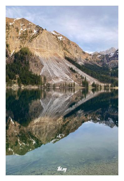 View across a mountain lake with a completely still mirror-like surface reflecting the mountains opposite, their rock faces illuminated by the warm orange evening sun. Small patches of snow near the summits. Semi-overcast sky.