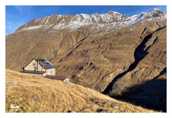 View of a mountain hut (Hochjoch Hospiz, already shuttered for the winter) in an empty, bare and vast high alpine landscape. The hut is seen from behind and from a distance, standing on a slope covered in dried brown grass, its color emphasized by the golden late afternoon light. Large mountains with snow-dusted peaks (incl. Kreuzspitze, Kreuzkogel, Sennkogel, Saykogel, all ~3300-3400m) are directly opposite with some deep canyons running down (carved out by the outflow of the melting glaciers)…