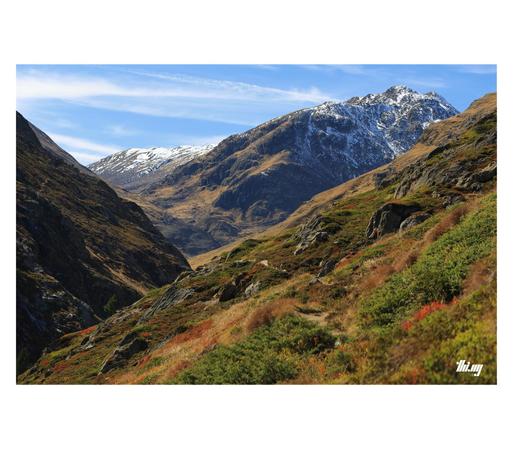 A wide and stunning mountain landscape with a small path leading through a dense colorful low-growth alpine vegetation (heather, bushes, blueberries, mosses) in different shades of browns, oranges, greens & reds. Gray rocks interspersed on the mountain slope which is leading down into a narrow canyon on the left-hand side. Large mountain peaks with a dusting of snow in the background. Pale blue sky with streaky clouds.