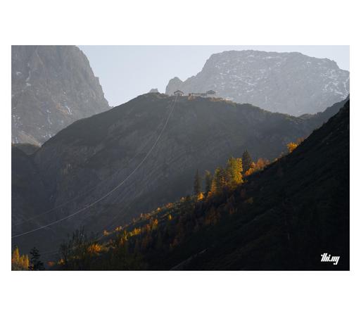 Multi-layered mountain landscape, in terms of distance, elevation and light. A mountain hut (Hanauer Hütte) is standing on the precipice of a large rock wall, the cables of its supply cable car leading up to it from the valley. Behind it, the ridge line of Schneekarlespitze and the north face of Dremelspitze in the haze. Most of the lower valley parts are already in deep shade from the surrounding peaks. Only a band of yellow/orange larches standing in the penumbra are still catching the beauti…