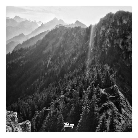High contrast black & white photo of an alpine mountain landscape and rugged ridge line in bright hazy evening light, seen from a summit cliff on said ridge. Incredible depth layering, god beams, lens flares. The slopes below the cliffs are covered in forest, in the background only visible as silhouettes.