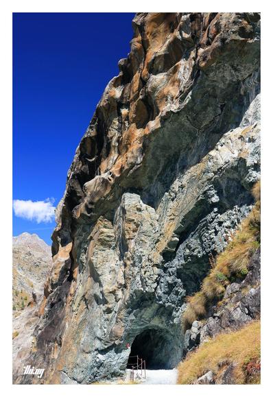 View of a small tunnel entrance (2-3 meters wide) into a glacially eroded colorful rock face consisting of slightly curved strata of different minerals and colors ranging from blueish-green grays to rusty orange. The higher up sections show signs of glacial plucking and trigger the imagination how that place must have looked filled with ice... A tall mountain peak in the far back. Clear, deep blue sky.