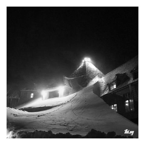Black and white night-time shot of the entrance area of Timberline Lodge with a huge snowdrift up until the top of the roof (equivalent of 4 floors). The sky is completely black and the scene is only lit by the big flood lights installed.