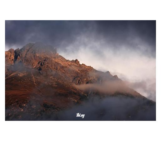 A mountain peak in rusty red/brown colors from the early morning alpen glow (and the general impact of October) against a pale beige sky and behind layers of dark gray clouds...