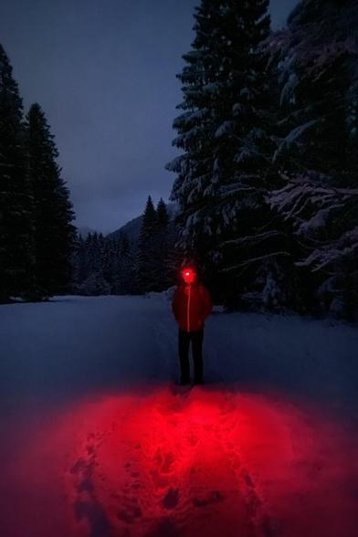 A person with a headlight standing on a faint path in approx. 30cm snow in a nighttime forest. The red headlight is casting a large HAL9000-stylee red glow in the snow in the foreground...