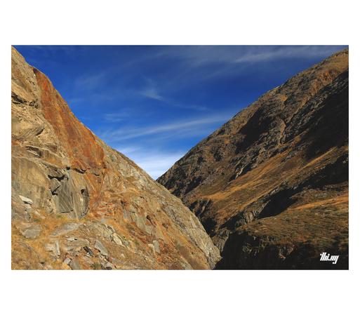 View of the upper slopes of a canyon in an empty high alpine landscape. The steep side on the left consists of mainly bare rocks (in colorful strata and shades of rust and ochre), also emphasized by the autumn light. A barely visible narrow path (partially secured via steel rope) is leading over the rocks into the distance. The other side of the canyon consists of rocks in rich dark browns, the lower slopes semi-covered in dry grasses, dropping off vertically into the deep shade near the bottom…