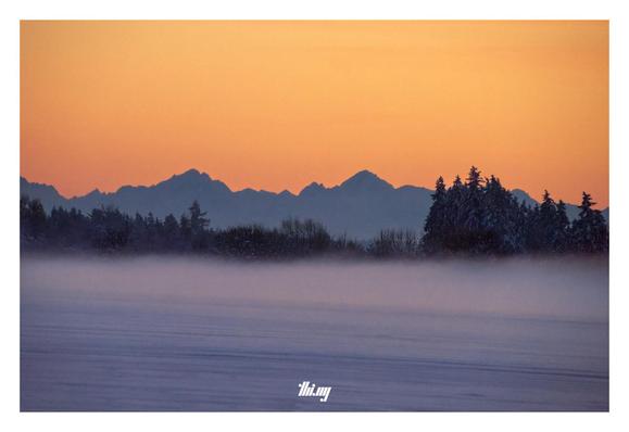 An empty winter landscape just after sunset, with frozen fog hovering over a snow plain in the foreground. Further back, the silhouettes of a stretch of snowy forest and behind that the silhouettes of the Northern Alps. Dominating it all is an intensely orange glowing sky.