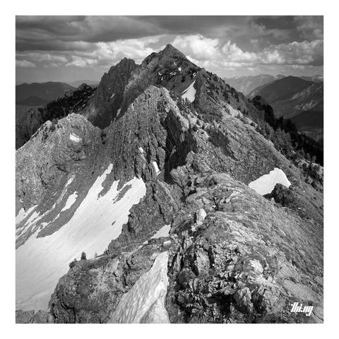 B&W photo of a very craggy ridge leading towards multiple peaks in the distance, with snow fields going down on the steep slopes on the left and more grassy slopes on the right. Parts of the forested neighboring valleys are visible in the distance. The main summit (Klammspitze) seems to almost touch the low hanging clouds whose shadows are also creating an interesting lighting, with only some parts of the ridge in bright sun...