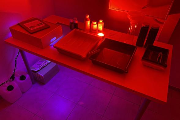 Long exposure photo of a basic DIY darkroom setup in my basement, featuring an old table with a bunch of photo trays (different sizes), bottles of chemicals for developing prints, a wooden box converted into an UV exposure unit for contact printing