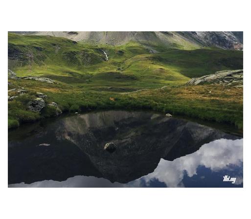 A small lake/pond in a high alpine landscape of only grasses and talus fields visible. The shallow water in the lake is very dark and reflecting some of the nearby mountain peaks of the surrounding 270 degree cirque. The framing of the image is such that only the reflection of the peaks and some of the clouds are visible.