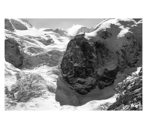 B&W photo of the near vertical section of a gigantic glacier, heavily crevassed and surrounded by large rock faces, heavily packed with snow. A cloud blowing over one of the summits in the back.