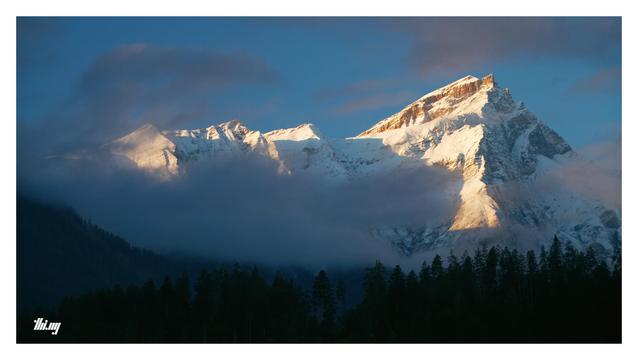 Cinematic wide-format view of a wintry snow covered mountain massif with its highest peak (Piz Beverin, 2998m) on the right side. The mountain is illuminated by golden morning light, some of the rocks glowing bright orange. In front are still partially dense low hanging clouds (formerly fog banks), but dispersing quickly and some of them with a faint pink tone. Below and further in the front, the silhouette of dense forest, still completely in deep shade. Bright blue sky.