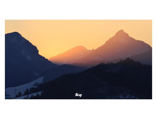 A mountainous winter landscape during sunset, the sky is completely filled with an intense, bright orange/pink glow. Silhouettes of mountains, both in the shade (in the foreground) and in/behind the glow.