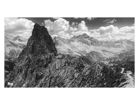 Black and white wide-format view of a narrow footpath near a mountain pass, going past a steep craggy cliff, before dropping a few hundred meters down (out of view). Far views into the mountains on the opposite side of the valley and beyond. An ancient gigantic side moraine on the opposite side of the valley... Semi cloudy sky.