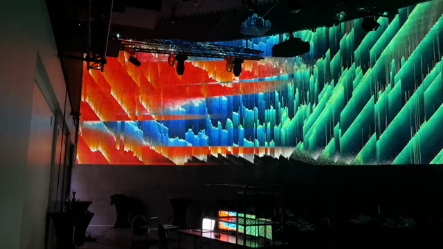Video panorama of a dark gallery space with a massive super-widescreen projection of an abstract, colorful 1.5D cellular automata artwork, with different automata creating different structures which are partially displacing and/or morphing with each other whilst they all co-evolve in the same shared space/environment.