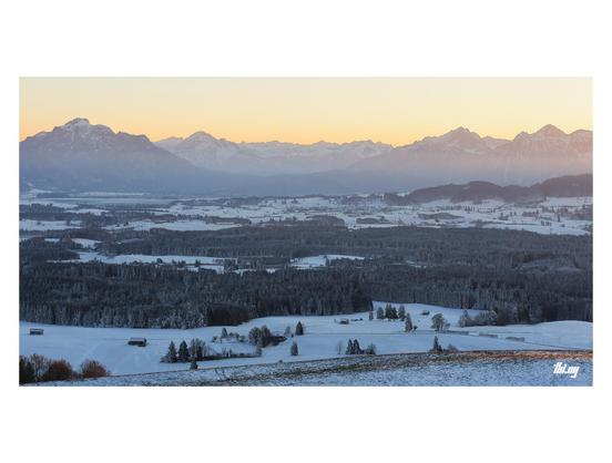 Wide-format photo of a beautiful snowy winter landscape of the foothills and peaks of the northern Alps near Füssen. The freezing temperatures and the setting sun is making the hazy sky glow orange/pink with cold blueish light in the lower region in the foreground. Partially snow-covered forest and fields with small wooden huts.