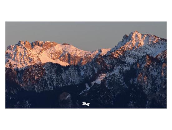 A long snowy mountain ridge (Hochplatte) and nearby peaks in the Ammergau Alps illuminated by the setting sun, glowing bright red/orange. The summit cross on the peak on the right (Branderschrofen) is glistening brightly. It's a freezing winter day and the steep lower slopes (partially forested) in the foreground are already in the shade (deep blueish light), only some cliffs are still reflecting the sun. Uniform gray clear sky.