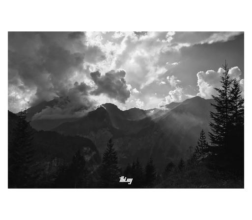 B&W photo of a mountain landscape in the Swiss Alps, with dramatic clouds forming and shafts of light reaching down into the deep valley. The silhouettes of trees in the foreground.
