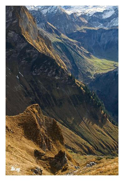 View of the upper section of Oytal and surrounding majestic mountains, as seen from a narrow hiking path near the ridge, a 1000 meters above the valley. Super steep grassy cliffs and slopes (in autumn colors) dropping down directly next to the path. Another small path winding up in the valley to the cirque in the distance. Some of these mountains there already dusted with snow.