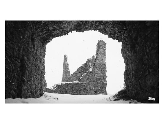 View of an old snowed in castle ruin with just a few remaining partial walls & tower structures, seen through (and framed by) the main gate. Plain white sky with large snow flakes.