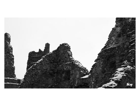 An old snowed in castle ruin with just a few remaining partial walls and tower structures. Plain white sky.