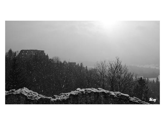 View of an old castle ruin standing on the top of the neighboring hill in a snow storm. A broken wall fragment of another castle in the foreground. In the distance some parts of the nearby valley visible, though limited visibility due to the snow, the fog and the sun trying to break through...